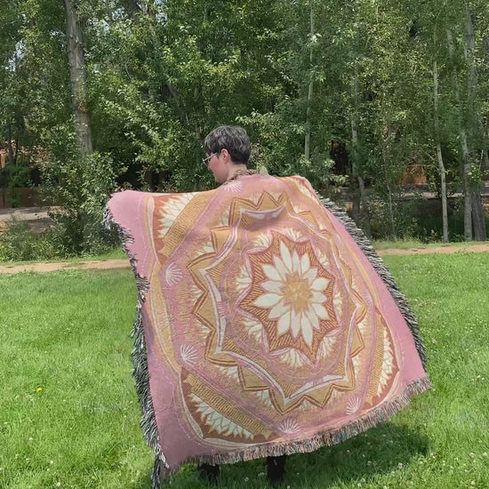 Mackenzie flips a pink and golden mandala woven blanket with fringe to see the front and back